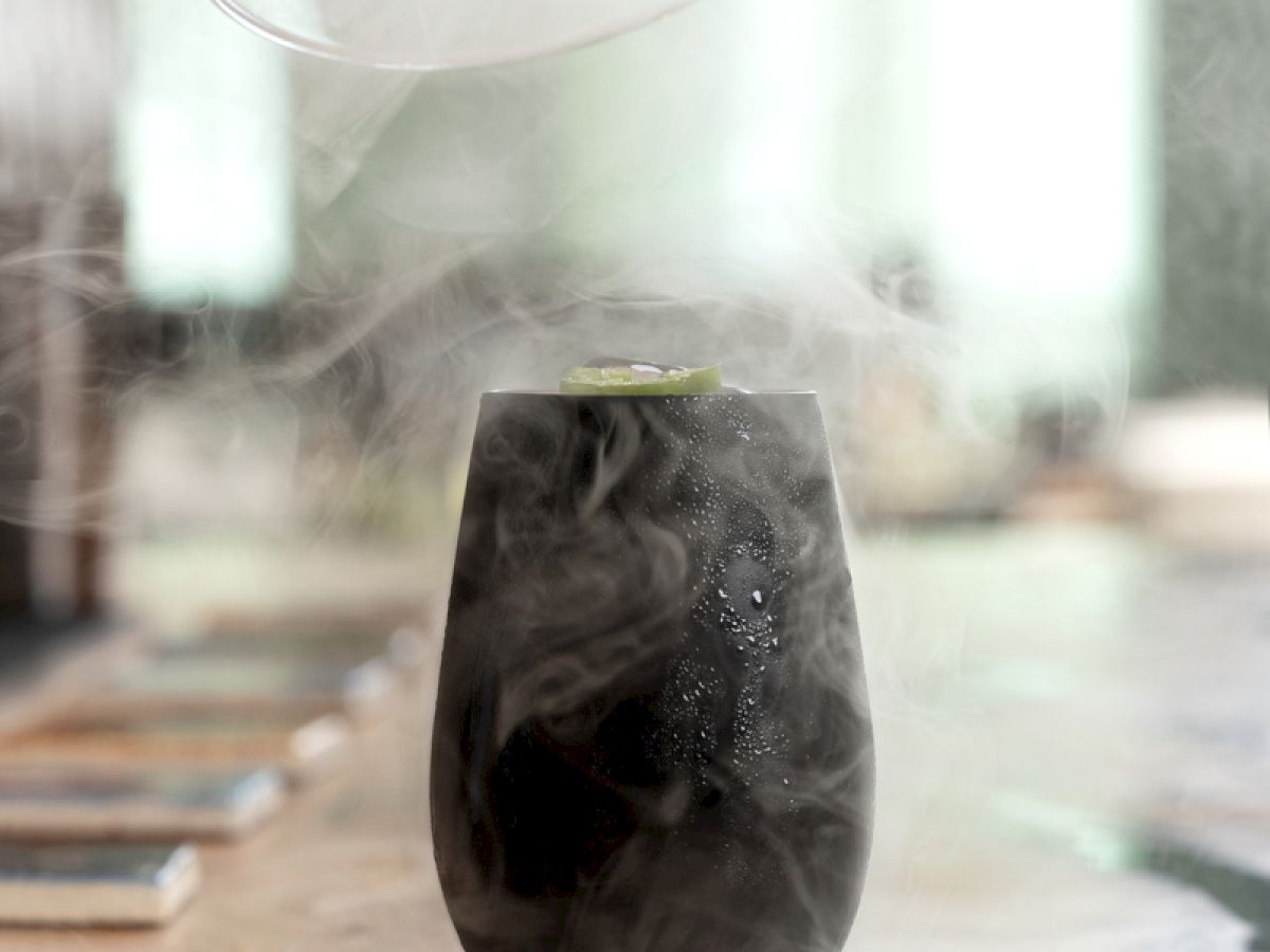 A smoke-filled glass is being uncovered, revealing a foggy vapor around it atop a wooden surface.
