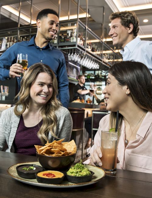 A group of people enjoying food and drinks in a lively restaurant setting, with appetizers and cocktails on the table.