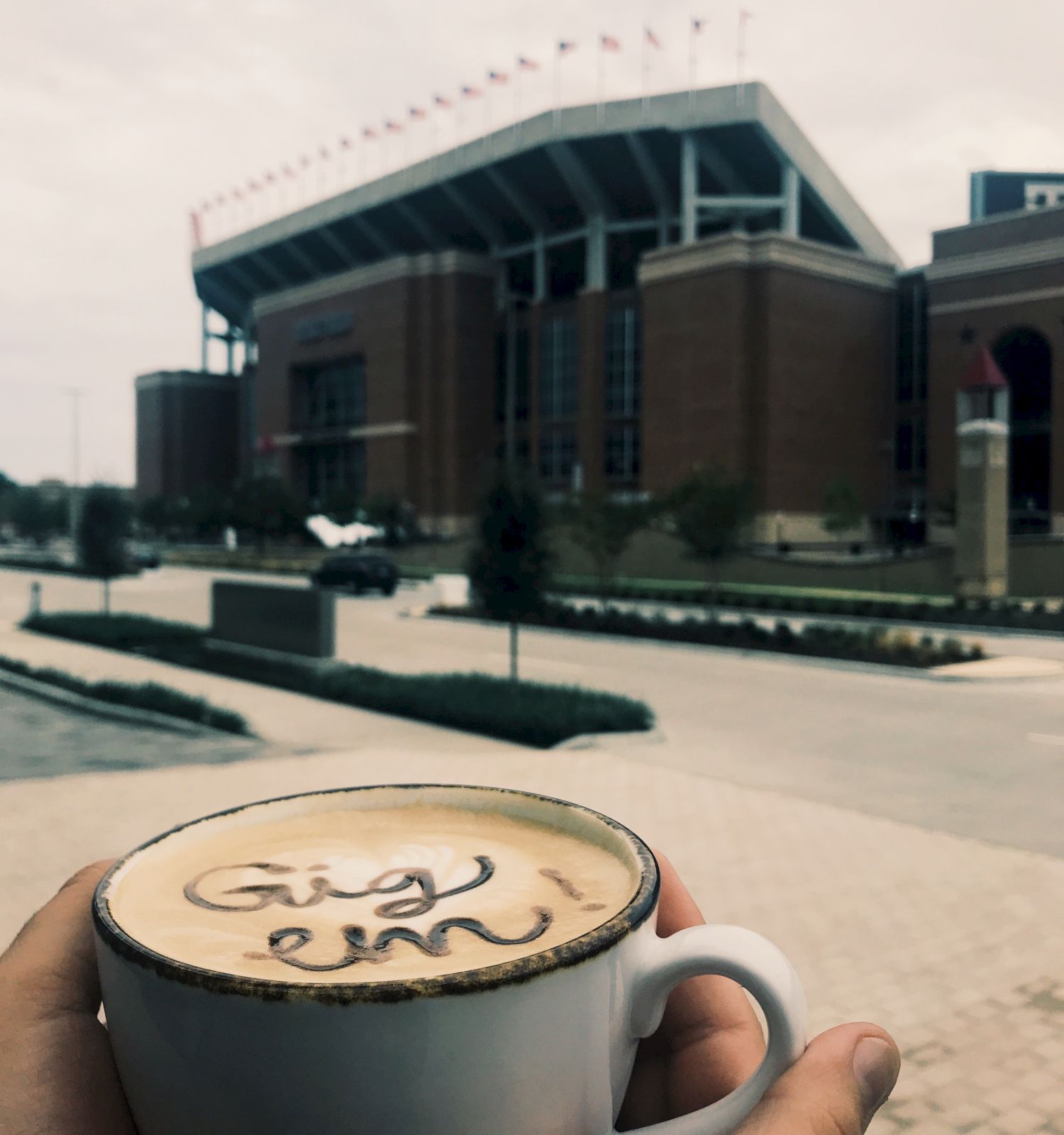A person holds a cup of coffee with "Gig 'em" written in the foam, in front of a large stadium.