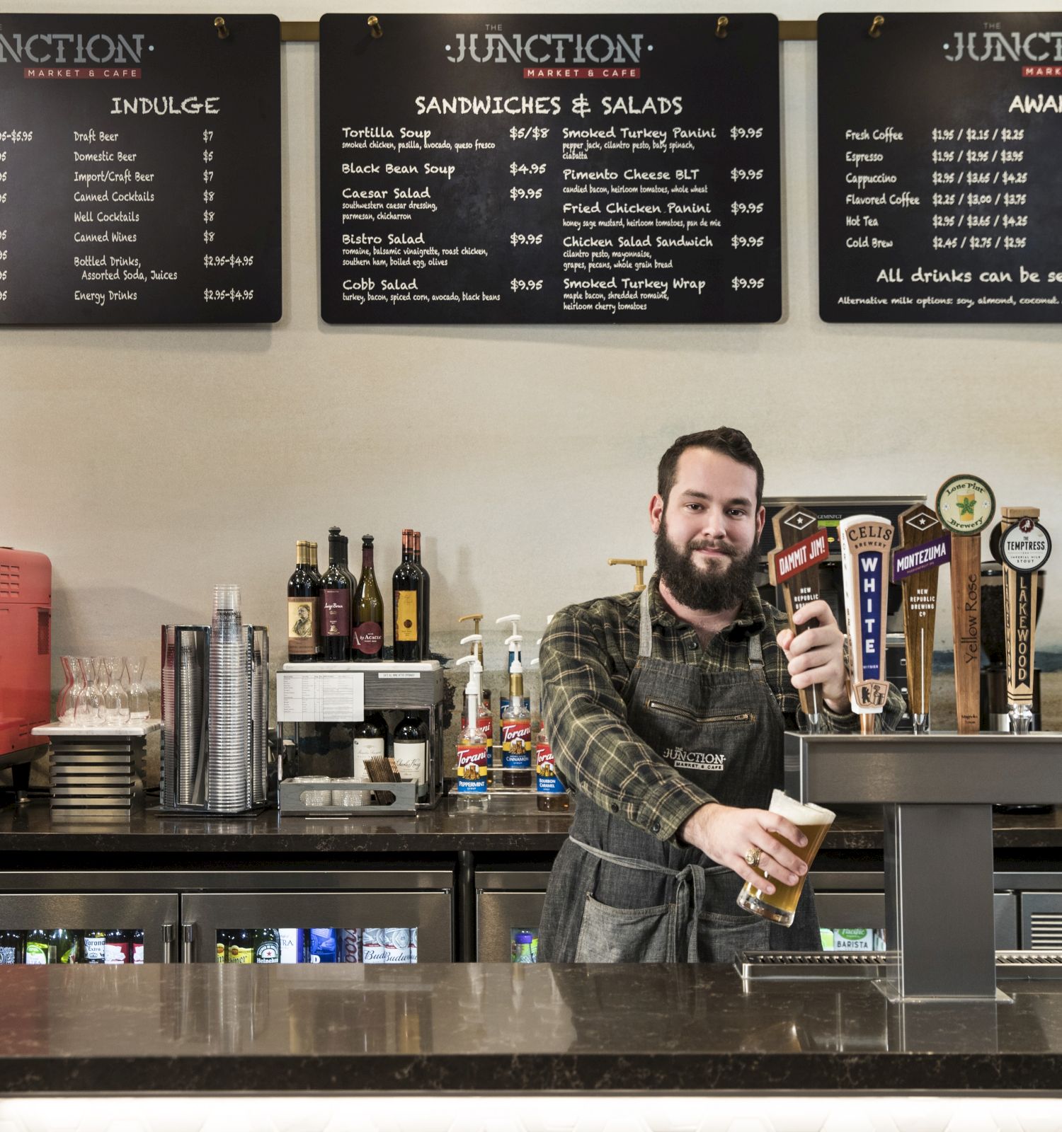 A bartender pours a draft drink behind a counter with a menu of sandwiches, salads, and drinks. Snacks, coffee machine, and taps visible.