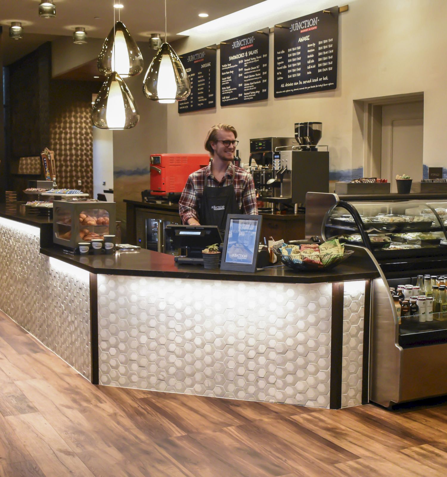 A coffee shop interior with a barista behind the counter, pastries, and beverages displayed. Modern decor and menu on the wall.