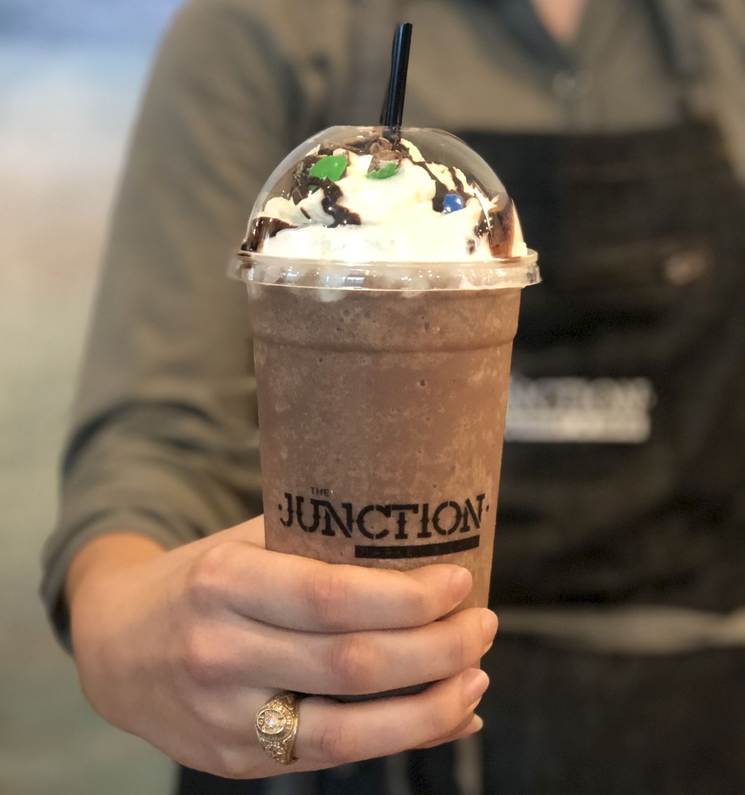 A person holding a frapp&eacute; with whipped cream and chocolate, in a plastic cup labeled "JUNCTION," topped with a domed lid and straw.