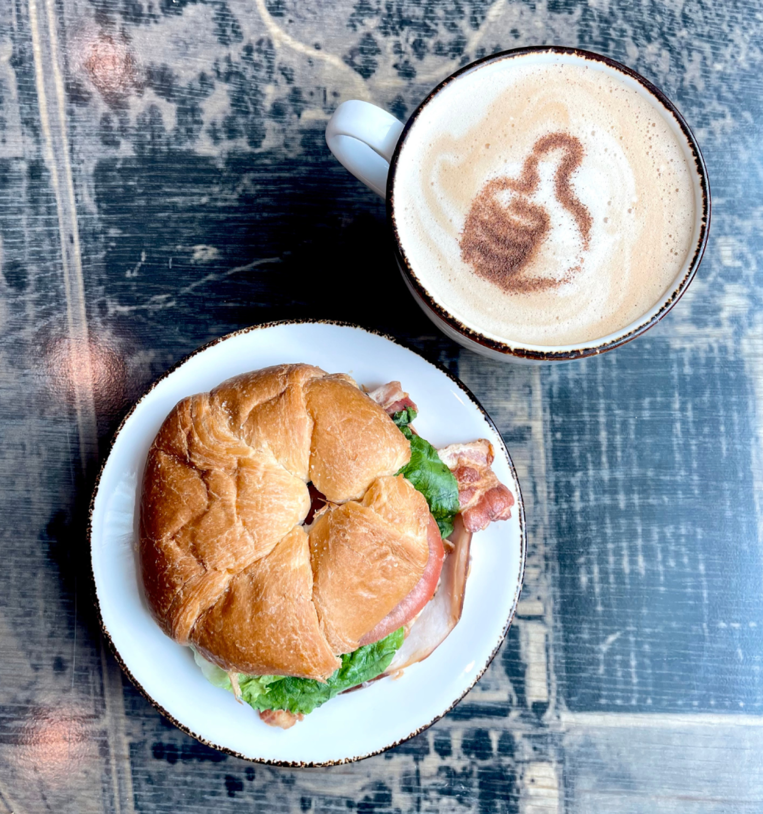 A croissant sandwich on a plate with lettuce and bacon is next to a cup of coffee with a design on the foam, placed on a rustic table.