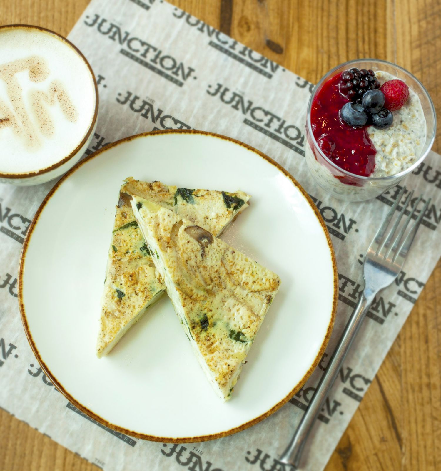 A plate with two slices of egg bread, a cappuccino with foam art, and a glass of chia pudding topped with berries on a wooden table.