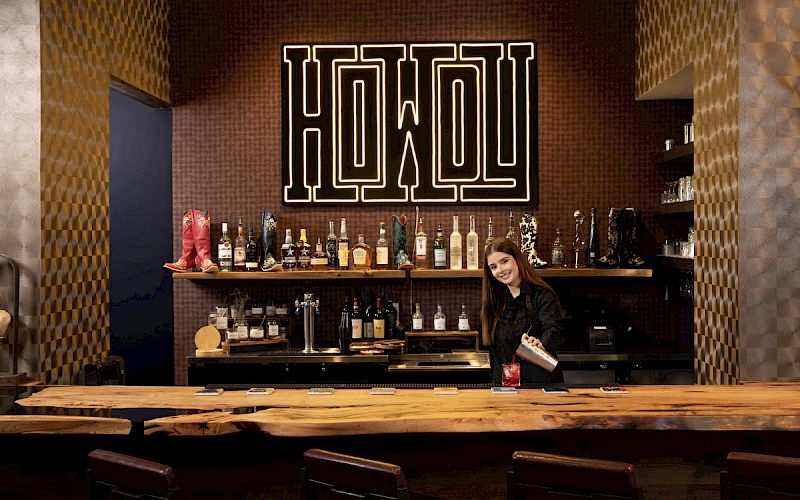 A bartender stands behind a well-stocked bar with a "Howdy" sign above. They're preparing a drink on a wooden counter in a cozy setting.
