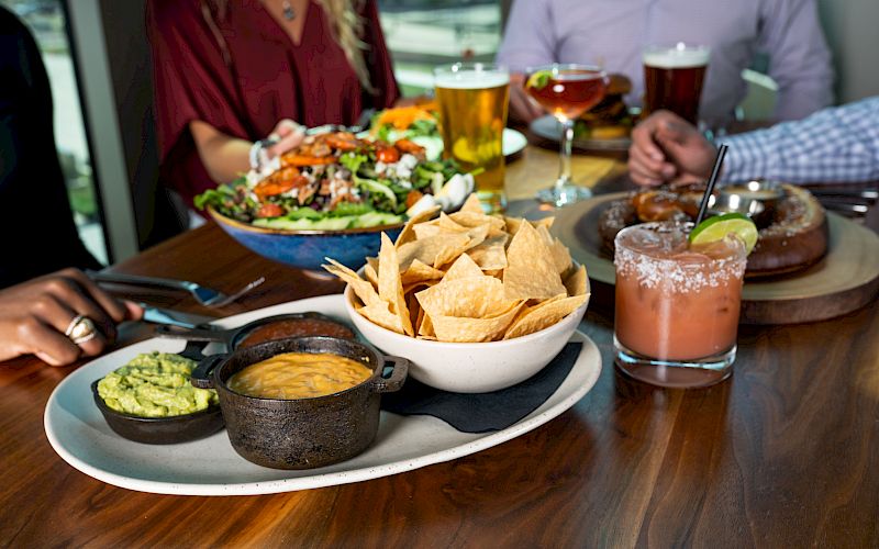 A table with tortilla chips, dips, a salad, and drinks. People are gathered, enjoying food and drinks together.