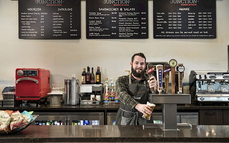 A bartender pours a beer at a counter with a menu overhead, surrounded by equipment and drinks, in a casual eatery setting.