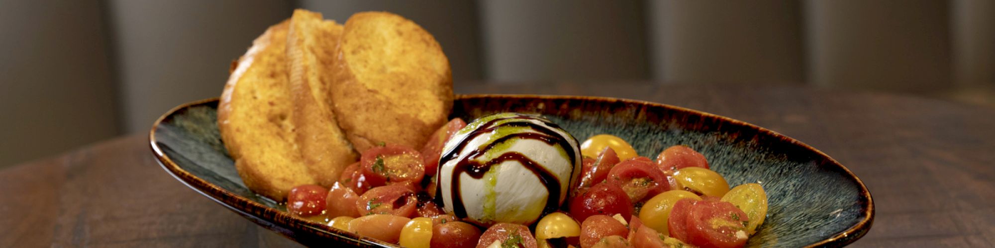 The image shows a plate with burrata cheese, cherry tomatoes, balsamic drizzle, and bread slices, placed on a wooden table.