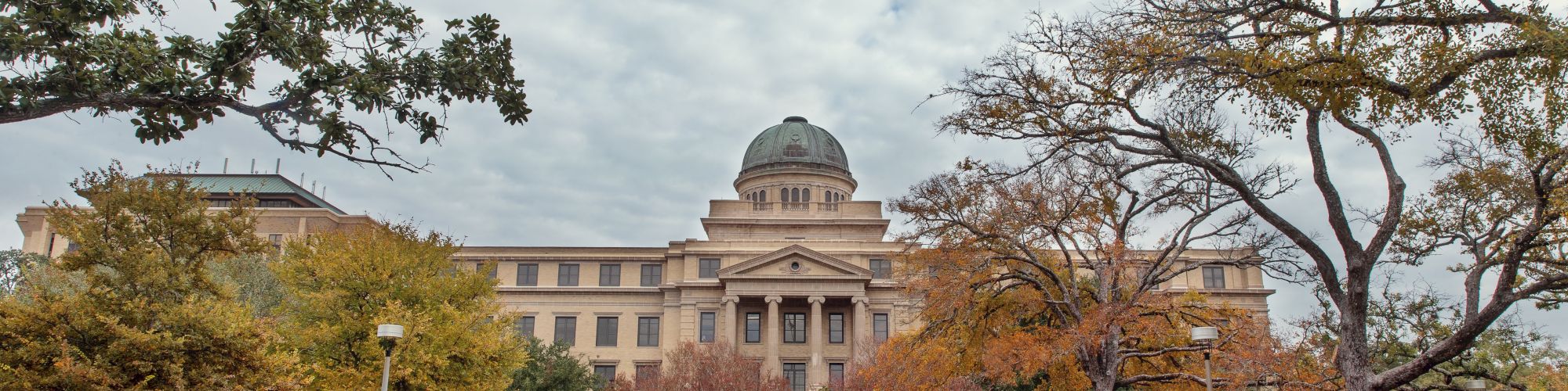 A university courtyard with a grand building and central dome, framed by autumn trees under a cloudy sky.