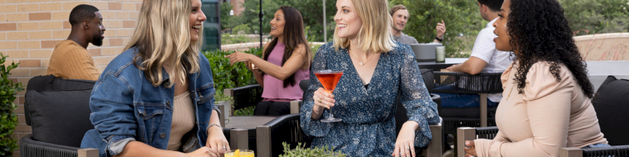 Drafting a sunny rooftop hangout: friends chat and sip colorful cocktails around a round table, with plants and red umbrellas in the background.