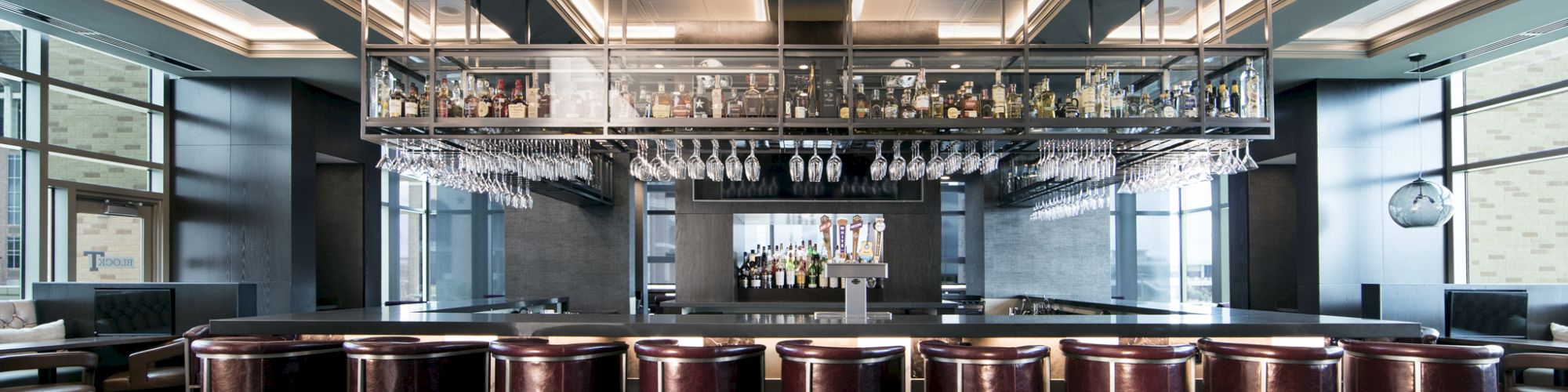 A modern bar interior with high stools, hanging glasses, and shelves of bottles. The space is sleek and well-lit with ambient lighting.