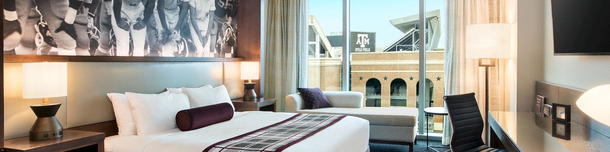 A hotel room with a large bed, art of football players, a desk, and a view of a stadium through a window is shown.