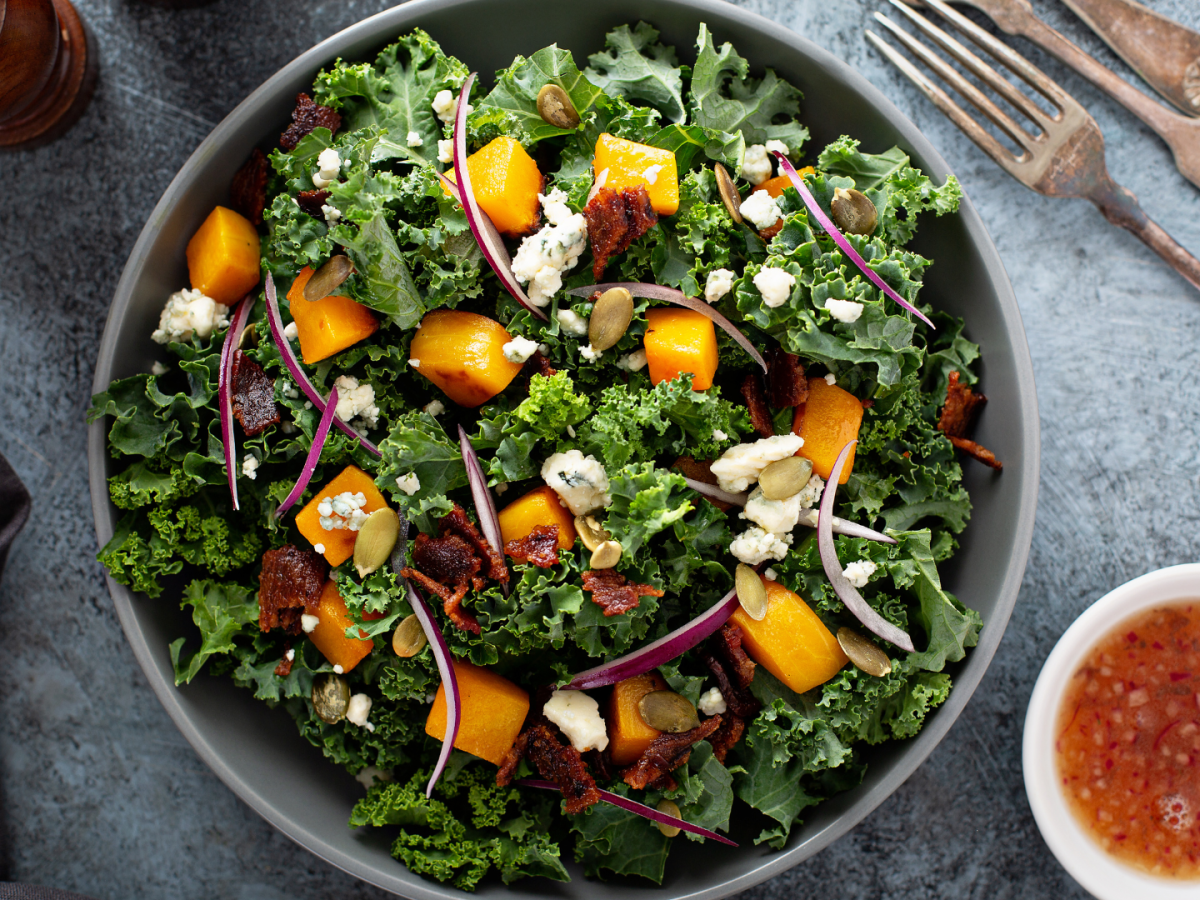 A kale salad with orange cubes, red onion slices, nuts, and cheese crumbles. On the side is a small bowl of dressing.