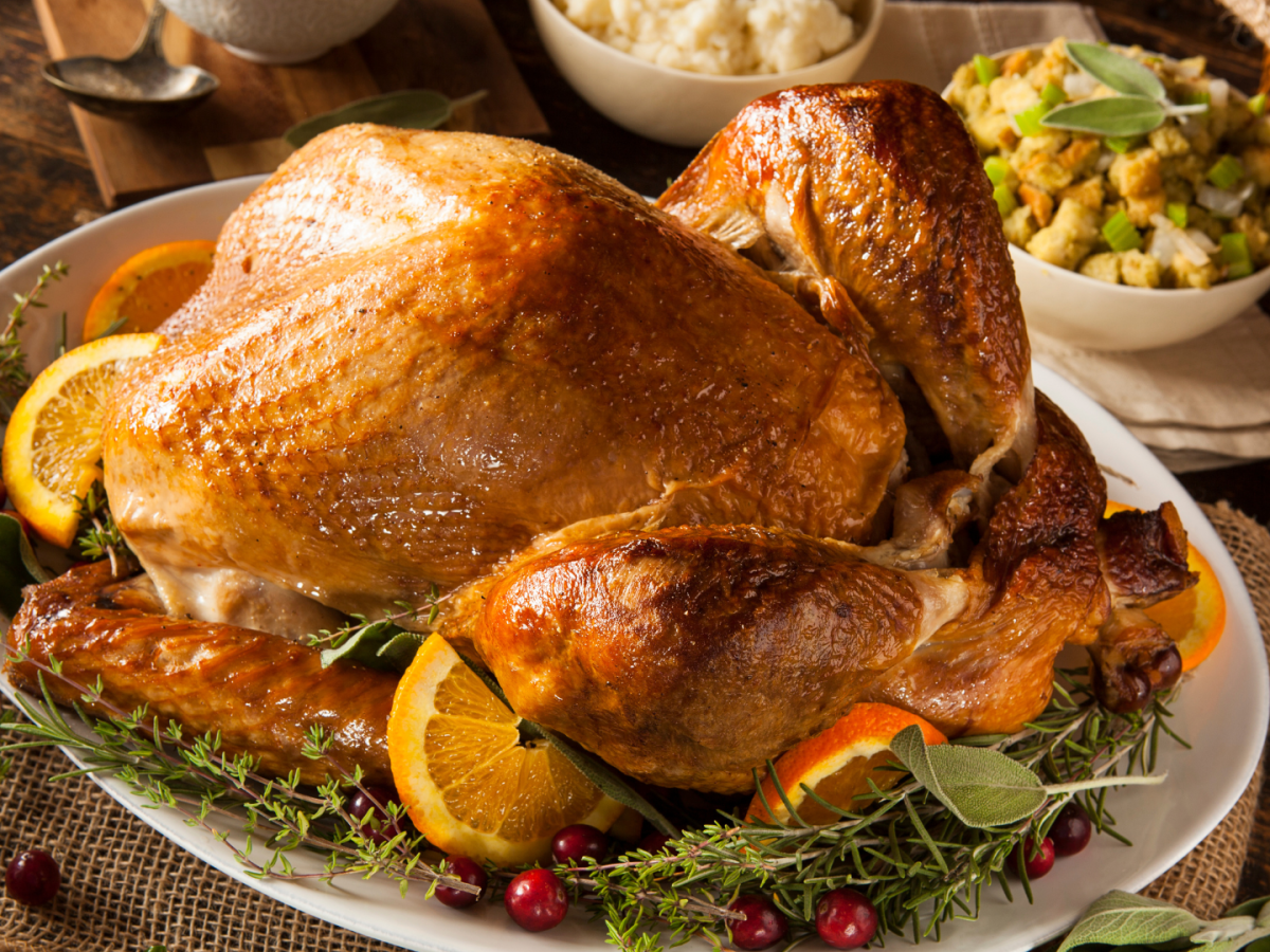A roasted turkey on a platter garnished with oranges and herbs, accompanied by stuffing and other side dishes on a wooden table.