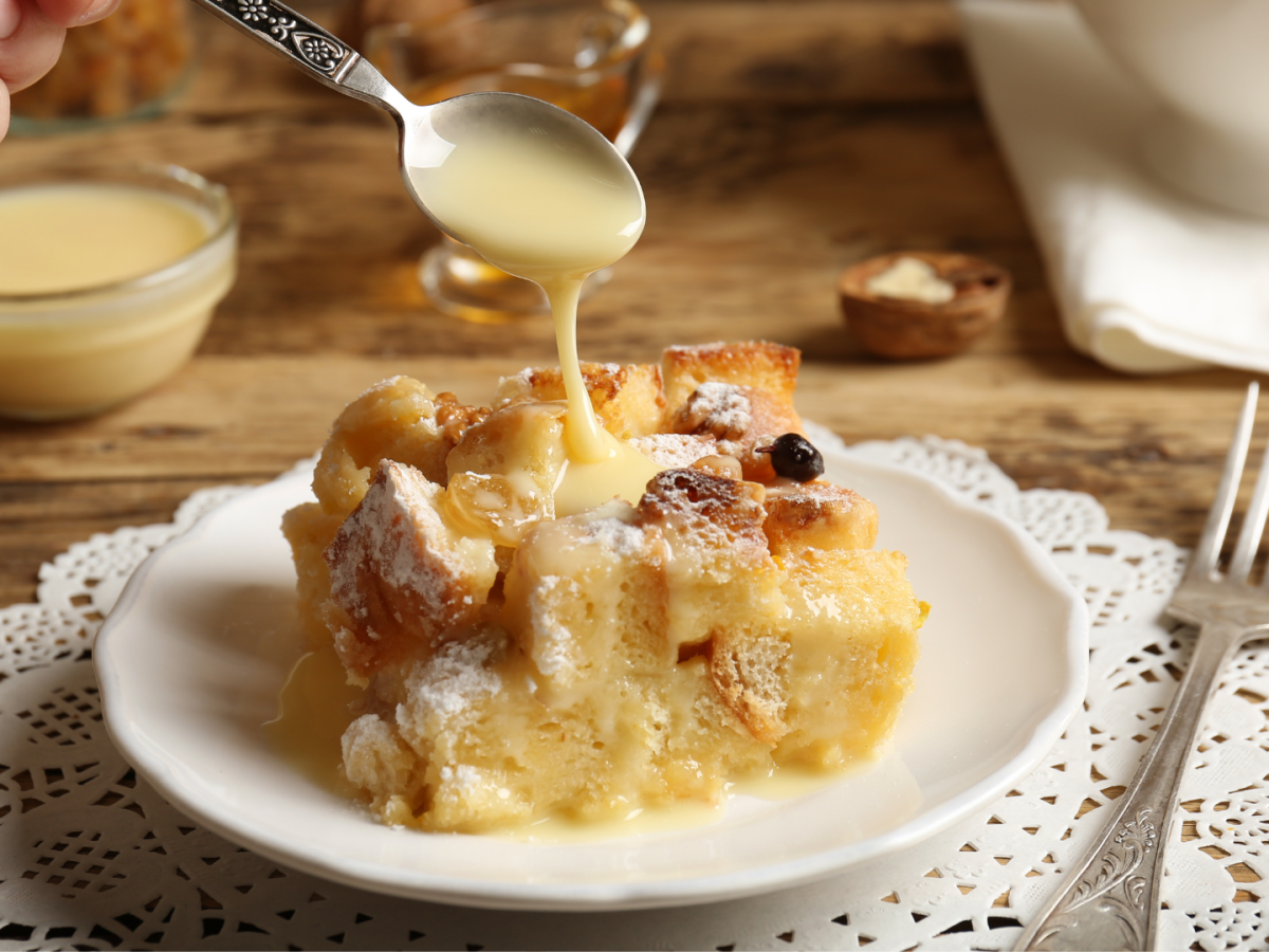 A plate of bread pudding topped with sauce is being served, accompanied by walnuts and cutlery on a wooden table with lace doily.