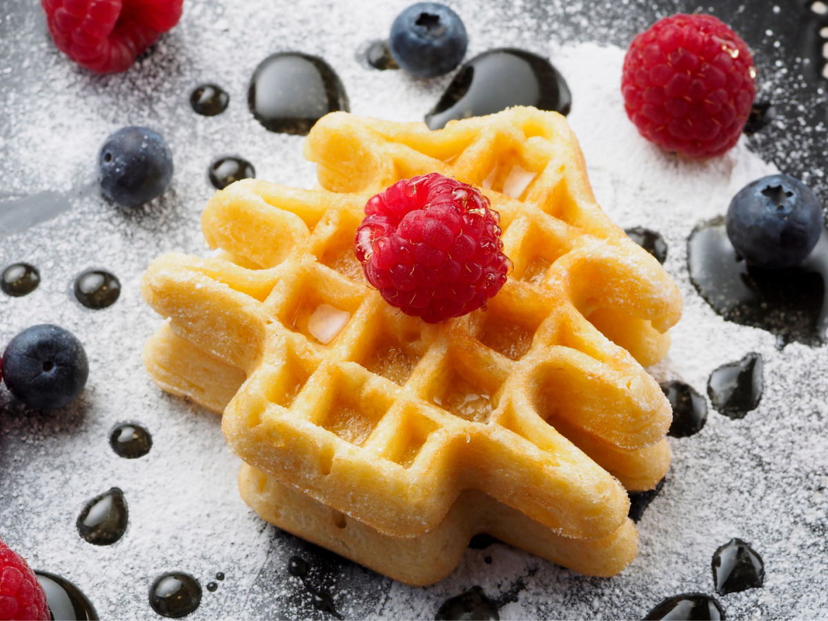 A waffle topped with raspberries, blueberries, powdered sugar, and syrup drops on a plate.