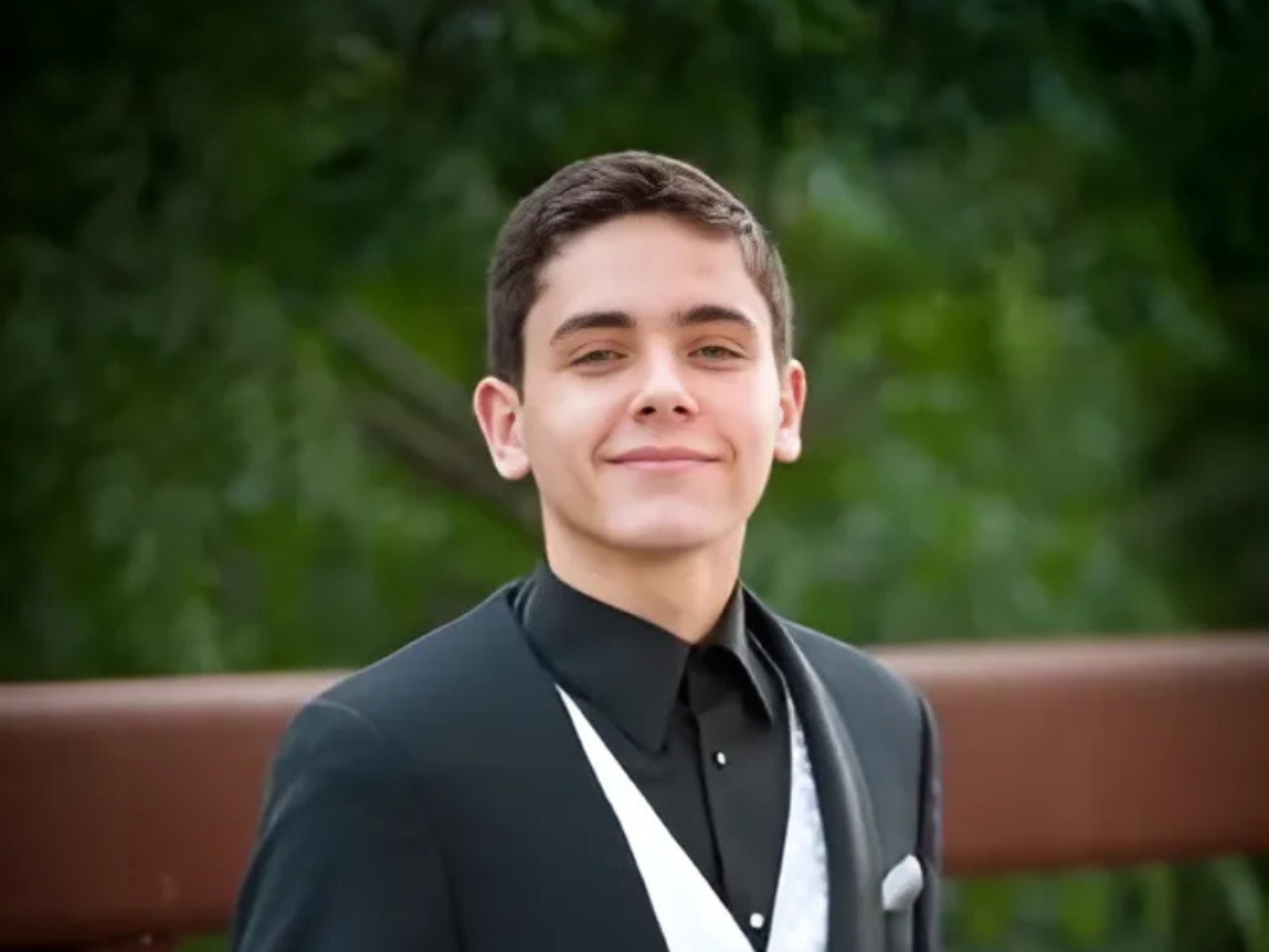 A person in a formal suit stands outdoors, with a wooden railing and greenery in the background.