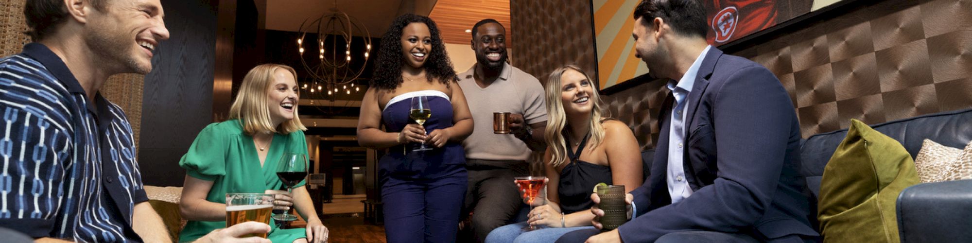 A group of people socializing in a modern lounge area with drinks, sitting near a painting of a person in a cowboy hat.