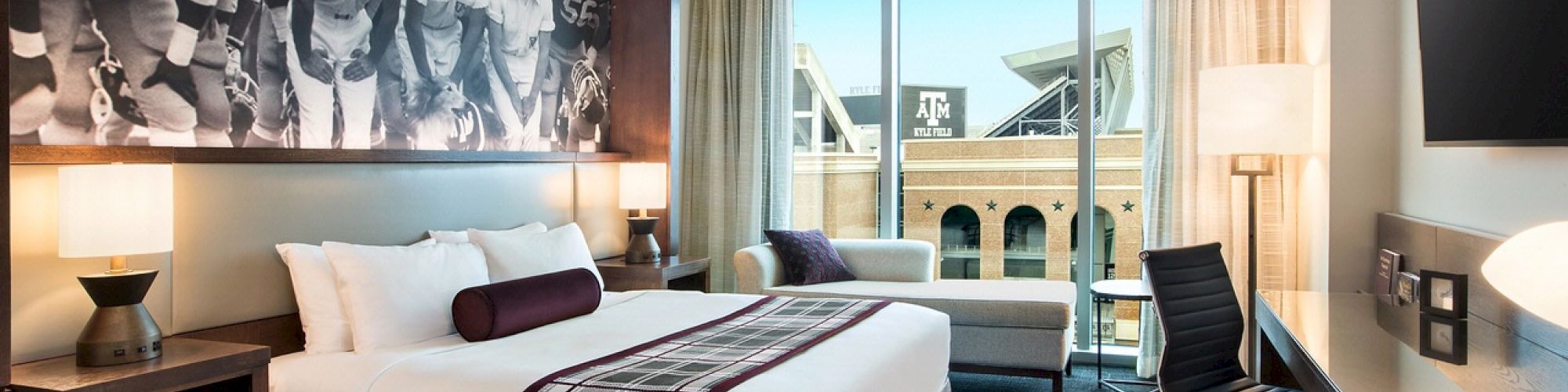 A modern hotel room featuring a tidy bed, Aggie-centric wall art, a desk and office chair, and grey lounging couch overlooking Kyle Field