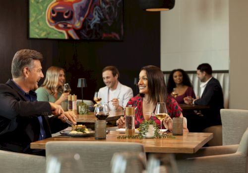 A group of adults dining at a restaurant, chatting and smiling around a wooden table with plates, drinks, and a cozy, social vibe.