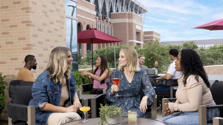 People sitting outdoors at a cafe, enjoying drinks and conversing by tables. There's a building in the background and a clear blue sky.