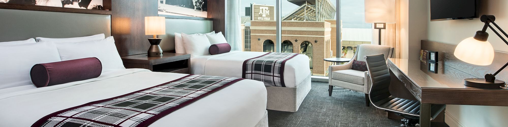 The image shows a modern hotel room with two beds, a large window view, wall art, a desk, chair, TV, and contemporary decor.