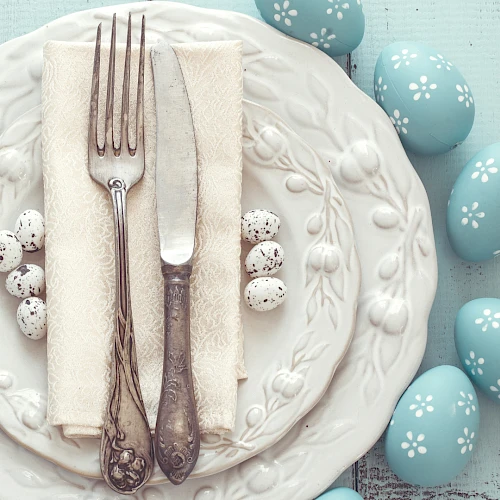 A neatly set plate with a folded napkin, fork, and knife, surrounded by blue Easter eggs on a light wooden table, with "Brazos" watermark.