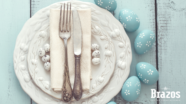 A neatly set plate with a folded napkin, fork, and knife, surrounded by blue Easter eggs on a light wooden table, with "Brazos" watermark.
