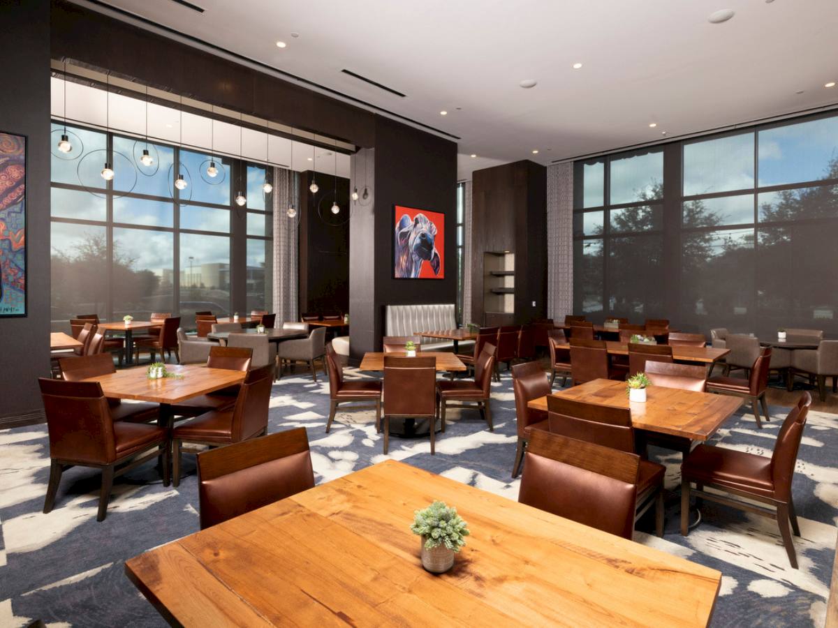 A modern, well-lit restaurant with wooden tables and chairs, large windows, mood lighting, artwork on the walls, and small potted plants on each table.