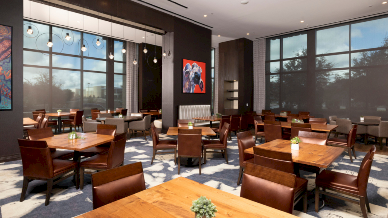 A modern, well-lit restaurant with wooden tables and chairs, large windows, mood lighting, artwork on the walls, and small potted plants on each table.