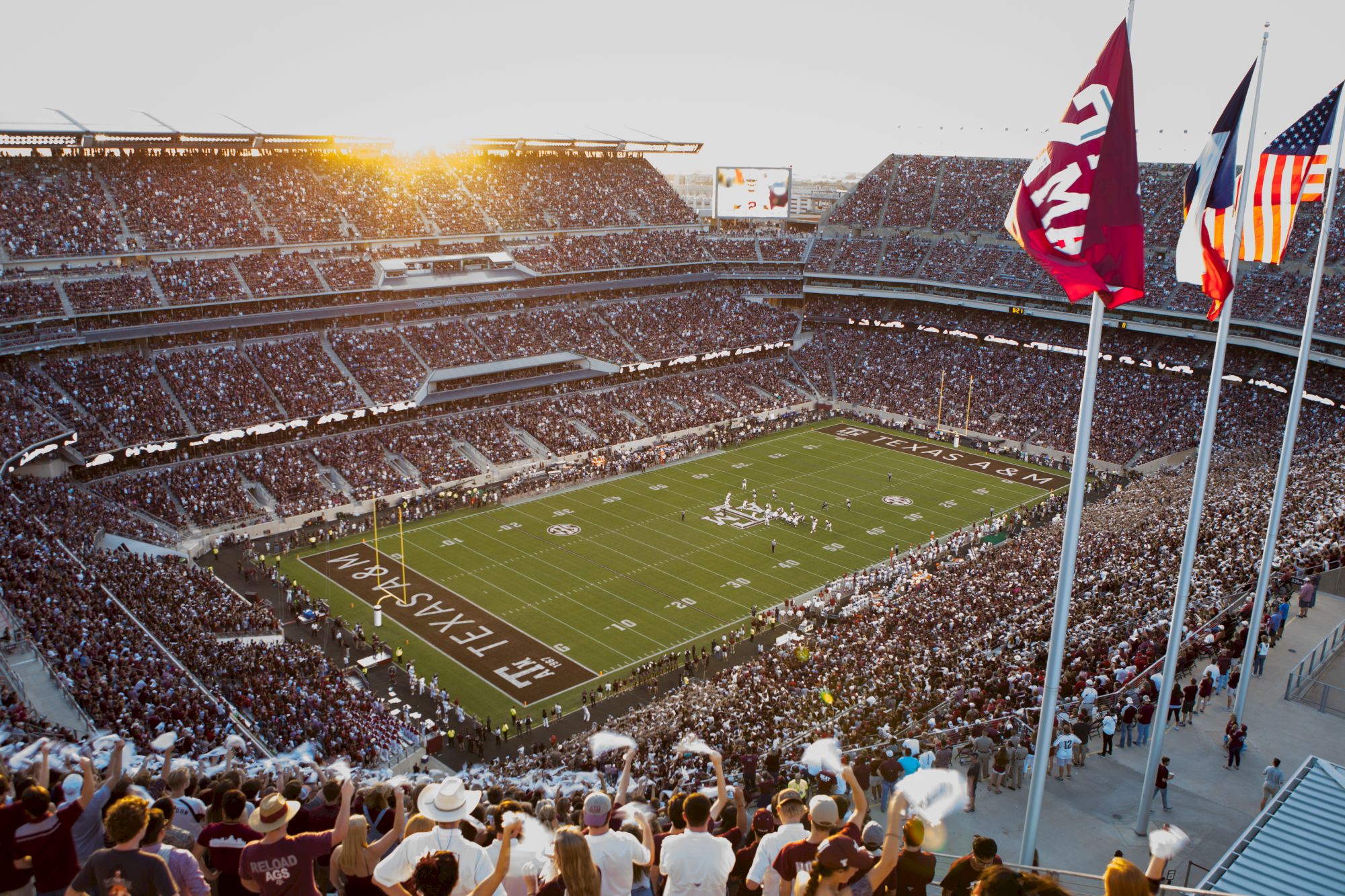 A crowded college football stadium at sunset, fans fill the stands, flags wave over a green field with players on the gridiron.