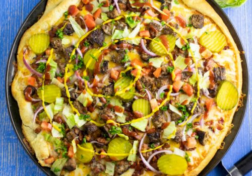 A loaded pizza with olives, peppers, onions, bacon bits, and greens on a crispy crust, served on a blue table with a knife nearby.
