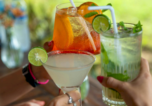 Three colorful cocktails clinked together in the foreground, each with garnishes, straws, and ice, set against a sunny outdoor backdrop.