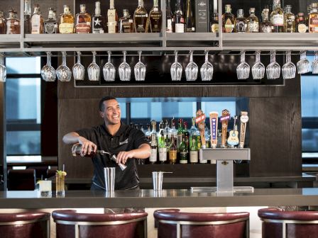 A bartender in a black shirt pouring drinks behind a bar, with bottles and glasses overhead and stools in front. The scene shows a lively bar setting.