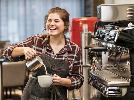 A barista smiles while pours milk into a cup under a coffee machine in a busy cafe.
